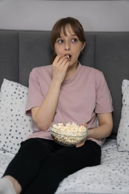 Şaşırmış genç bir kadın bir kase patlamış mısırla yatakta oturuyor, rahat yatak odasında, yumuşak bir ışık ve rahat bir atmosferle çevrili, bir film veya televizyon programının keyfini çıkarıyor. Dikey fotoğraf