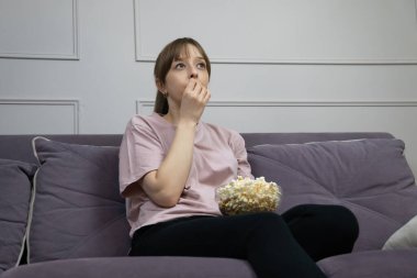 Sıkıcı genç kadın rahat bir kanepede oturmuş, elinde patlamış mısırla evde sıkıcı bir televizyon programı izliyor, boş zamanlarında ilgisizliğini ve yorgunluğunu dile getiriyor.