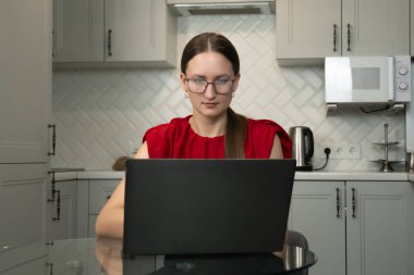 Gözlüklü ve kırmızı gömlekli genç bir kadın modern bir mutfakta cam bir masada oturuyor, bir dizüstü bilgisayarla işine odaklanıyor, muhtemelen uzaktan çalışıyor veya ev ekonomisini yönetiyor.