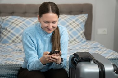 Akıllı telefon kullanırken gülümseyen genç bir kadın, paketlenmiş bir bavulun yanında bir yatakta oturuyor, yaklaşan bir iş gezisine ya da tatil macerasına hazırlanıyor.