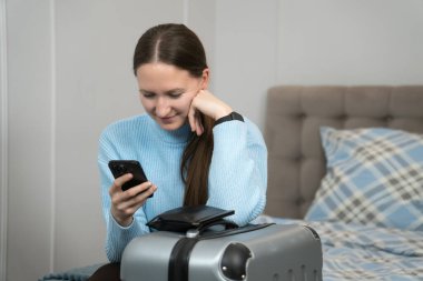 Bir otel odasında bavulun üzerinde oturan genç bir turist seyahat uygulamalarını kontrol etmek için akıllı telefonunu kullanıyor ve heyecan verici bir gezi ve macera planlarını tamamlıyor.