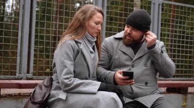 Genç bir adam ve kadın parkta bir bankta oturuyor, kulaklıklarını paylaşıyor, cep telefonundan müzik dinliyor, serin bir sonbahar gününde yürüyüş sırasında bir anlık bağlantının tadını çıkarıyorlar.