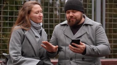 Genç bir adam ve kadın parkta bir bankta oturuyor, kulaklıklarını paylaşıyor, cep telefonundan müzik dinliyor, serin bir sonbahar gününde yürüyüş sırasında bir anlık bağlantının tadını çıkarıyorlar.