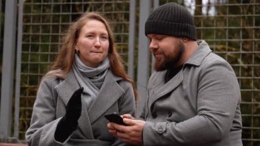 Genç bir adam ve kadın parkta bir bankta oturuyor, kulaklıklarını paylaşıyor, cep telefonundan müzik dinliyor, serin bir sonbahar gününde yürüyüş sırasında bir anlık bağlantının tadını çıkarıyorlar.