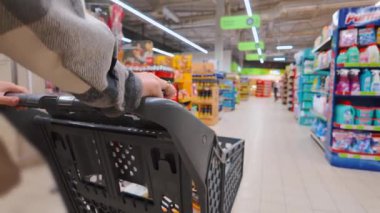 Bir kadının el arabasının, arka planda çeşitli ürünlerle dolu raflarla dolu parlak bir market koridorunda rehberlik edişinin birinci şahıs görüntüsü.
