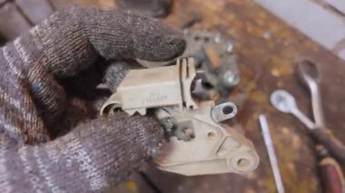 Profesyonel oto tamircisi koruyucu eldivenler giyer ve bir aracın alternatör voltaj düzenleyici ünitesini kontrol eder, bir tamirhanedeki fırçaları ve genel durumu kontrol eder.