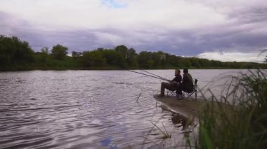 Erkek ve kadın birlikte kaliteli zaman geçiriyorlar bulutlu bir günde, nehrin kumlu kıyısında çubuklarla balık avlıyorlar. Açık hava hobilerinin, boş zamanlarının ve sakin bir hafta sonu inzivasının tadını çıkarıyorlar.