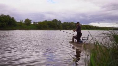 Erkek ve kadın birlikte kaliteli zaman geçiriyorlar bulutlu bir günde, nehrin kumlu kıyısında çubuklarla balık avlıyorlar. Açık hava hobilerinin, boş zamanlarının ve sakin bir hafta sonu inzivasının tadını çıkarıyorlar.