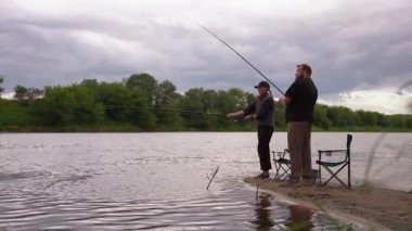 Erkek ve kadın birlikte kaliteli zaman geçiriyorlar bulutlu bir günde, nehrin kumlu kıyısında çubuklarla balık avlıyorlar. Açık hava hobilerinin, boş zamanlarının ve sakin bir hafta sonu inzivasının tadını çıkarıyorlar.