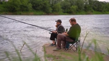 Erkek ve kadın birlikte kaliteli zaman geçiriyorlar bulutlu bir günde, nehrin kumlu kıyısında çubuklarla balık avlıyorlar. Açık hava hobilerinin, boş zamanlarının ve sakin bir hafta sonu inzivasının tadını çıkarıyorlar.