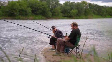 Erkek ve kadın birlikte kaliteli zaman geçiriyorlar bulutlu bir günde, nehrin kumlu kıyısında çubuklarla balık avlıyorlar. Açık hava hobilerinin, boş zamanlarının ve sakin bir hafta sonu inzivasının tadını çıkarıyorlar.