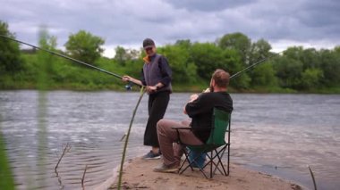 Genç çift, kumlu bir nehir kıyısında balık avlama hobilerinin keyfini çıkarırken, kadın ayakta duruyor ve erkek sakin bir yaz gününde oltalarını tutarken bir kamp sandalyesinde oturuyor.