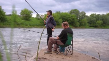 Genç çift, kumlu bir nehir kıyısında balık avlama hobilerinin keyfini çıkarırken, kadın ayakta duruyor ve erkek sakin bir yaz gününde oltalarını tutarken bir kamp sandalyesinde oturuyor.