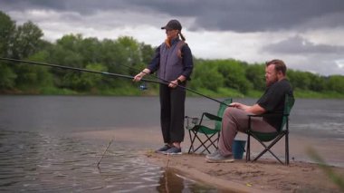 Erkek ve kadın birlikte kaliteli zaman geçiriyorlar bulutlu bir günde, nehrin kumlu kıyısında çubuklarla balık avlıyorlar. Açık hava hobilerinin, boş zamanlarının ve sakin bir hafta sonu inzivasının tadını çıkarıyorlar.