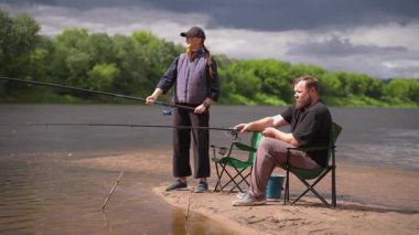 Genç çift, kumlu bir nehir kıyısında balık avlama hobilerinin keyfini çıkarırken, kadın ayakta duruyor ve erkek sakin bir yaz gününde oltalarını tutarken bir kamp sandalyesinde oturuyor.