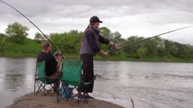 Sakin bir adam ve kadın nehir kenarındaki katlanan sandalyelerde oturur, sabırla olta kamışı tutar ve bir ısırık bekler, bulutlu bir günde birlikte sakin bir açık hava hobisinin keyfini çıkarırlar.