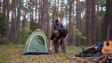 Beyaz bir erkek ve kadın, güzel bir çam ormanında yeşil bir çadır kurmak için birlikte çalışıyorlar. Kamp yerlerini, yakınlarda bir gitar ve kamp malzemesi olan açık hava macerasına hazırlıyorlar.