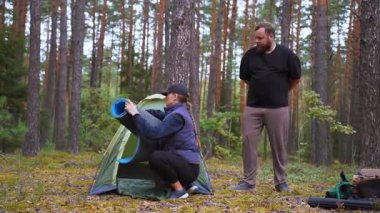 Genç çift pitoresk bir çam ormanında kamp kurarken, eşi yakınlarda durup süreci izlerken, kadın yeşil bir çadırda bir uyku paspası düzenliyor.