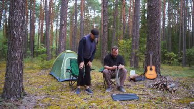 Genç bir adam ve kadın güzel bir çam ormanında kamp alanı kuruyorlar. Kamp sandalyelerini açıyorlar ve hafta sonu kaçamağı için çadırlarının yanında taşınabilir küçük bir masa var.