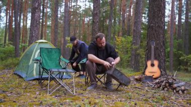 Genç çift, kamplarını güzel bir çam ormanına kuruyor. Kadın dışarıda geçirecekleri macera için çadır ve ekipmanları hazırlarken, adam portatif barbekü ızgarası yapıyor.