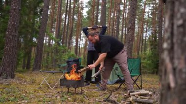 Genç bir kadın kamp ateşinin yanında esneme egzersizleri yaparken bir adam da kamp alanının yakınında oturuyor. Aktif yaşam tarzı ve arka planda çadır ve şenlik ateşi olan açık hava eğlencesi.