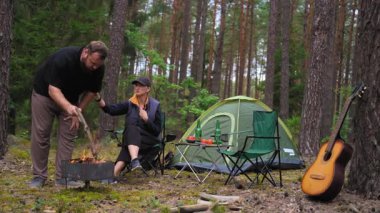 Genç çift ormanda huzurlu bir akşamın tadını çıkarırken kadın çadırlarının yakınındaki bir sandalyede dinlenirken erkek şenlik ateşiyle ilgilenir ve rahat ve samimi bir ortam yaratır.