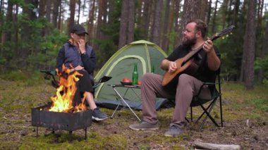 Genç bir çift orman kamp alanında romantik bir akşamın tadını çıkarırken, bir kadın arka planda çadırıyla birlikte sıcak bir şenlik ateşinin yanında şarkı söylerken sakallı bir adam gitar çalıyor.