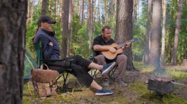 Genç bir çift, kamp alanında sakin bir akşamın tadını çıkarıyor. Sakallı bir adam bir kadın için akustik gitar çalıyor. Kamp sandalyelerinde çadırın yanında dinleniyor ve kamp ateşi yakıyor.