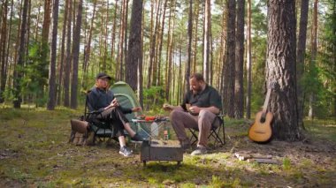 Genç bir çift çadırın yanında katlanan sandalyelerde oturuyor, barbekü pikniğinin tadını çıkarıyor ve güzel bir çam ormanında rahatlatıcı bir hafta sonu kaçamağı sırasında sohbet ediyorlar.