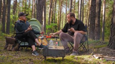 Genç bir çift çadırın yanında katlanan sandalyelerde oturuyor, barbekü pikniğinin tadını çıkarıyor ve güzel bir çam ormanında rahatlatıcı bir hafta sonu kaçamağı sırasında sohbet ediyorlar.