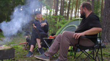 Ormandaki kamp sandalyelerinde oturan genç bir çift çadırın yanında ızgara yemek yiyip sohbet ediyorlar. Kadın sohbet sırasında el kol hareketi yaparken adam şişeden bira içiyor.
