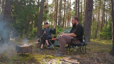Genç bir çift çadırın yanında katlanan sandalyelerde oturuyor, barbekü pikniğinin tadını çıkarıyor ve güzel bir çam ormanında rahatlatıcı bir hafta sonu kaçamağı sırasında sohbet ediyorlar.