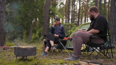 Ormandaki kamp sandalyelerinde oturan genç bir çift çadırın yanında ızgara yemek yiyip sohbet ediyorlar. Kadın sohbet sırasında el kol hareketi yaparken adam şişeden bira içiyor.