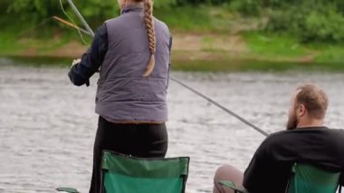Arkaya bakan bir adam ve kız, bulutlu bir günde arka planda yemyeşil ağaçlarla birlikte kumlu nehir kıyısındaki katlanan sandalyelerde birlikte balığın tadını çıkarıyorlar.