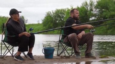 Kadın ve erkek birlikte sakin bir gün geçirmenin tadını çıkarıyorlar. Nehir kenarındaki katlanır sandalyelerde oturuyorlar. Çubukları suyun içinde.
