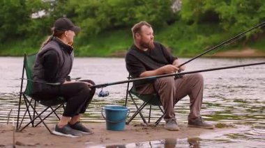 Bir erkek ve bir kadın, birlikte huzurlu bir gün geçirmenin tadını çıkarıyorlar. Nehir kenarındaki sandalyelerde oturuyorlar. Çubukları suya dökülmüş, yemyeşil ağaçlar ve sakin doğayla çevrili.