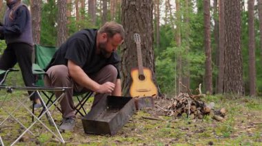 Genç çift, kamplarını güzel bir çam ormanına kuruyor. Kadın dışarıda geçirecekleri macera için çadır ve ekipmanları hazırlarken, adam portatif barbekü ızgarası yapıyor.