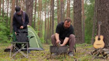 Genç çift, kamplarını güzel bir çam ormanına kuruyor. Kadın dışarıda geçirecekleri macera için çadır ve ekipmanları hazırlarken, adam portatif barbekü ızgarası yapıyor.