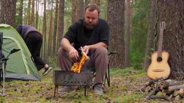 Genç çift, kamplarını güzel bir çam ormanına kuruyor. Kadın dışarıda geçirecekleri macera için çadır ve ekipmanları hazırlarken, adam portatif barbekü ızgarası yapıyor.