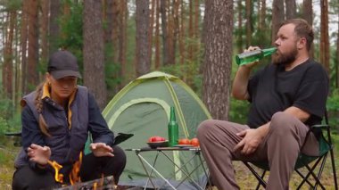 Genç bir çift ateşin yanında kamp sandalyelerinde oturur, yeşil şişelerden bira içerler ve konuşurlar, ve arkalarında güzel bir çam ormanı ortamında çadır ve piknik masası vardır.