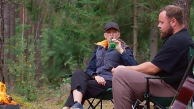 Genç çift sessiz bir kaçamağın tadını çıkarıyor, kamp sandalyelerinde oturuyor, bir şişeden bira içiyor ve sakin bir çam ormanında, bir sonbahar akşamı, sıcak bir kamp ateşinin yanında konuşuyor.