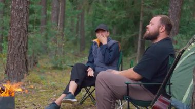 Genç bir çift ateşin yanında kamp sandalyelerinde oturur, yeşil şişelerden bira içerler ve konuşurlar, ve arkalarında güzel bir çam ormanı ortamında çadır ve piknik masası vardır.