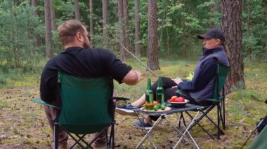 Genç bir çift ateşin yanında kamp sandalyelerinde oturur, yeşil şişelerden bira içerler ve konuşurlar, ve arkalarında güzel bir çam ormanı ortamında çadır ve piknik masası vardır.
