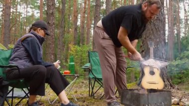 Sakallı bir adam kömür ızgarasında et pişirirken partneri de çam ormanındaki huzurlu bir piknik sırasında bir sandalyede rahatlarken doğa ve açık hava eğlencesinin tadını çıkarıyor.
