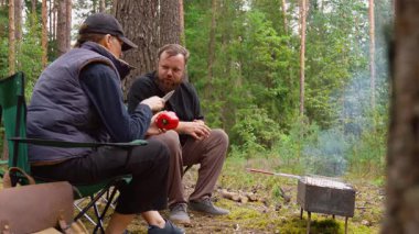 Sakallı bir adam kömür ızgarasında et pişirirken partneri de çam ormanındaki huzurlu bir piknik sırasında bir sandalyede rahatlarken doğa ve açık hava eğlencesinin tadını çıkarıyor.