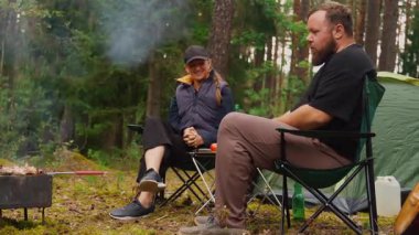 Mutlu genç çift ormanda kamp sandalyelerinde oturuyor, konuşuyor ve gülüyor, barbekü pikniğinde et pişiriyor ve temiz hava alıyor ve bira içiyor.