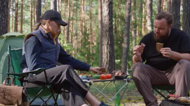 Genç bir çift güneşli bir ormanda iskemlede oturuyor, piknik sırasında ızgara et ve sebze yiyor, doğanın tadını çıkarıyor ve birlikte basit bir yemek yiyorlar.