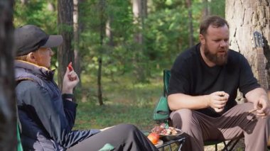 Genç bir çift güneşli bir ormanda iskemlede oturuyor, piknik sırasında ızgara et ve sebze yiyor, doğanın tadını çıkarıyor ve birlikte basit bir yemek yiyorlar.