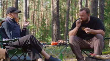 Genç bir çift güneşli bir ormanda iskemlede oturuyor, piknik sırasında ızgara et ve sebze yiyor, doğanın tadını çıkarıyor ve birlikte basit bir yemek yiyorlar.
