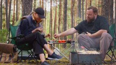 Genç adam ve kadın katlanır sandalyelerde oturur, yemek yer ve konuşurlar. Kamp alanlarında çadır ve çam ormanlarında ızgara tüttürerek rahatlatıcı bir pikniğin tadını çıkarırlar.